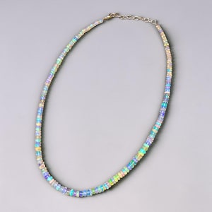 May include: A delicate necklace composed of small, iridescent opal beads. The beads display a range of colors, including blue, green, yellow, and pink, strung together with a silver chain. The necklace is displayed against a gray background.