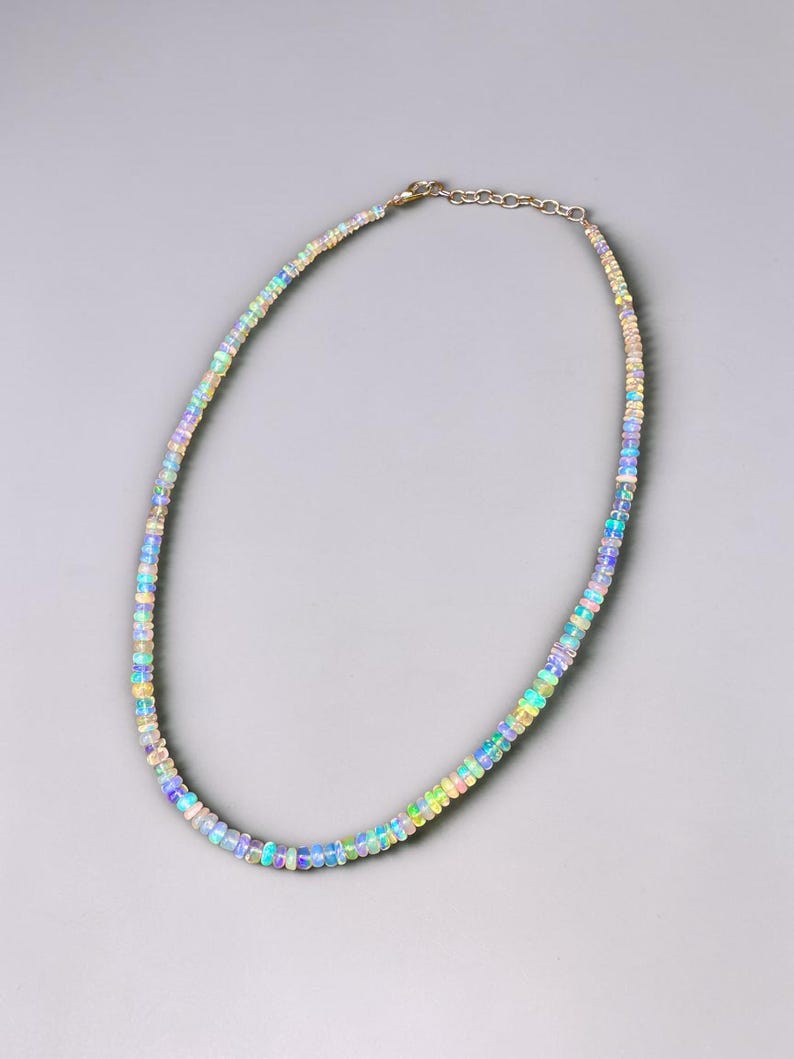 May include: A delicate necklace composed of small, iridescent opal beads. The beads display a range of colors, including blue, green, yellow, and pink, strung together with a silver chain. The necklace is displayed against a gray background.
