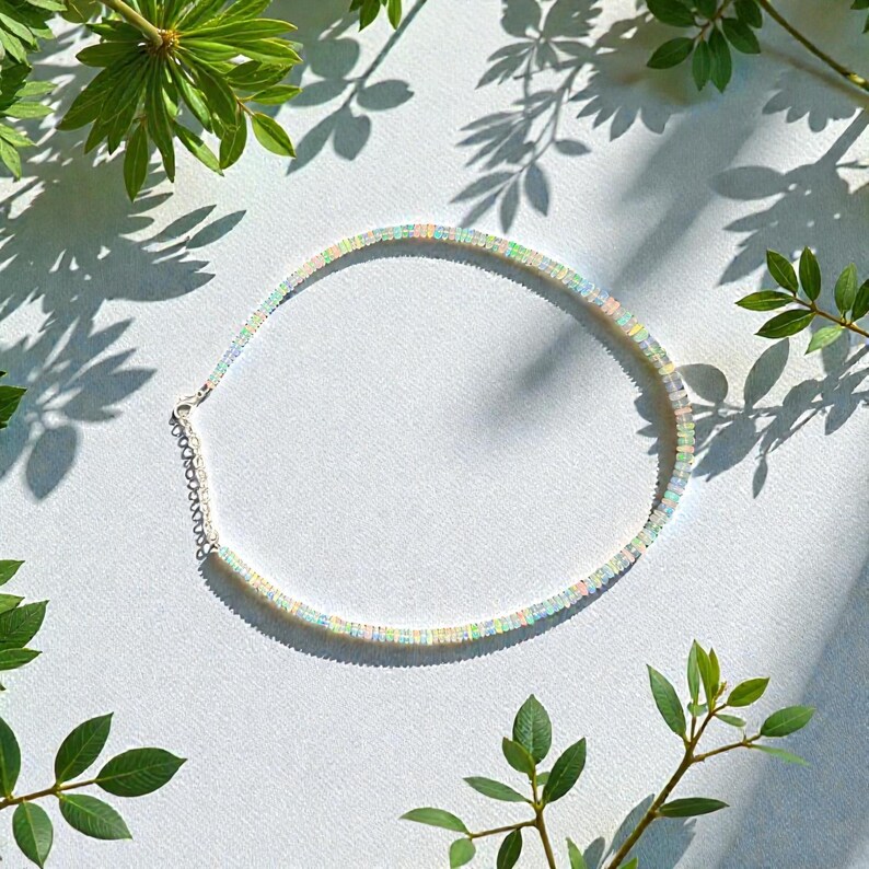 May include: A delicate opal necklace with a silver clasp and chain, resting on a white surface. The necklace is surrounded by green leaves and their shadows, creating a natural and elegant presentation. The opal beads display a range of colors.