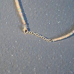 May include: Close-up of a delicate necklace featuring small, iridescent beads in shades of white, peach, and pale green. The necklace has a gold-colored chain and clasp, with an adjustable length. The jewelry is set against a textured gray background.