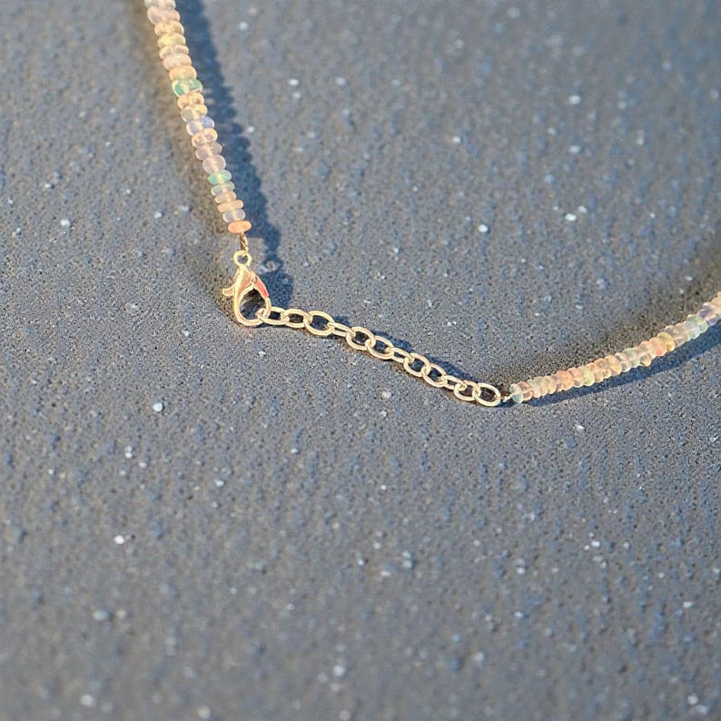 May include: Close-up of a delicate necklace featuring small, iridescent beads in shades of white, peach, and pale green. The necklace has a gold-colored chain and clasp, with an adjustable length. The jewelry is set against a textured gray background.