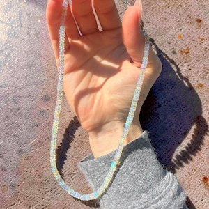 May include: A delicate necklace composed of small, iridescent opal beads. The necklace is strung on a silver chain and is held up against a neutral background. The opal beads display a range of colors, including white, yellow, and blue.