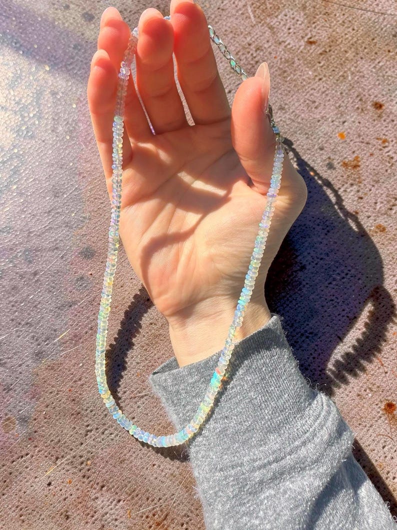 May include: A delicate necklace composed of small, iridescent opal beads. The necklace is strung on a silver chain and is held up against a neutral background. The opal beads display a range of colors, including white, yellow, and blue.