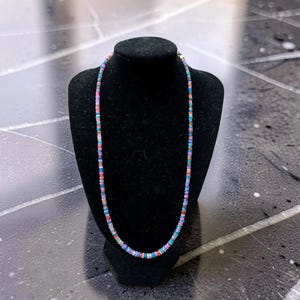 Puede incluir: Un collar compuesto por pequeñas cuentas circulares multicolores en tonos azul, rosa, blanco y naranja. Las cuentas están ensartadas en un patrón repetitivo para formar una sola hebra que cuelga en forma circular alrededor del expositor.