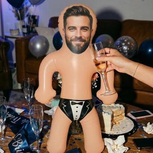 Könnte beinhalten: Aufblasbare Puppe mit Männergesicht, trägt einen Badeanzug im Smoking-Stil. Die Puppe steht auf einem Tisch mit Sektgläsern, Kuchen und einem "Bride Tribe"-Schärpe. Ballons im Hintergrund.