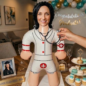 May include: An inflatable doll in a nurse's uniform, featuring a photo of a woman in a graduation cap. The doll has a stethoscope and is surrounded by celebratory items, including cupcakes and macarons. A banner reads "Congratulations Graduate!"