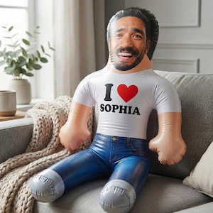 Könnte beinhalten: Aufblasbare Puppe mit lächelndem Gesicht, trägt ein weißes T-Shirt mit dem Aufdruck "I ❤️ SOPHIA" und eine blaue Jeans. Die Puppe sitzt auf einem grauen Sofa.