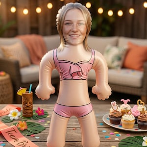 Puede incluir: Muñeca inflable con la cara de una mujer con un bikini rosa. La muñeca está sobre una mesa de madera con una banda "Team Bride", un vaso tiki y cupcakes. El fondo incluye un sofá y luces de cadena.