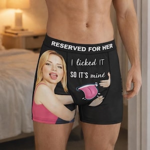 May include: Black boxer shorts with the text "RESERVED FOR HER" and "I licked IT so it's mine". The shorts feature a graphic of a woman and a tongue. The person is standing in a room.