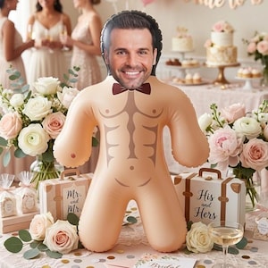 Pode incluir: Uma boneca insuflável com rosto masculino e torso musculoso, usando uma gravata borboleta. A boneca é bege e está rodeada de decorações de casamento, incluindo flores, bolos e caixas de presente. A cena sugere um chá de panela.