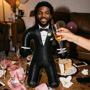 May include: An inflatable doll dressed in a black tuxedo, featuring a smiling face, bow tie, and a flower. The doll is surrounded by party decorations, including balloons, champagne glasses, and a cake. A pink sash with "Bride Tribe" is also visible.