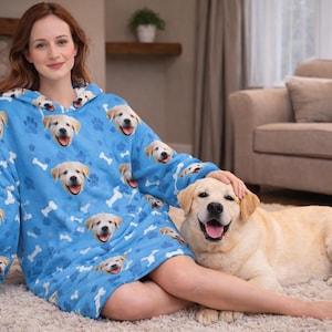 Puede incluir: Una manta con capucha azul claro con un patr&oacute;n repetido de caras de golden retriever sonrientes y huesos blancos. Un perro golden retriever est&aacute; acostado en una alfombra junto a la manta.