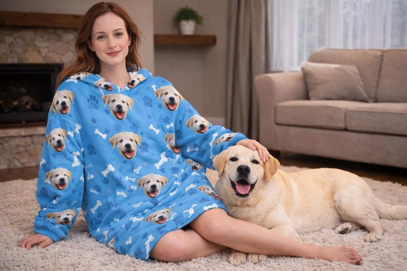 Puede incluir: Una manta con capucha azul claro con un patr&oacute;n repetido de caras de golden retriever sonrientes y huesos blancos. Un perro golden retriever est&aacute; acostado en una alfombra junto a la manta.