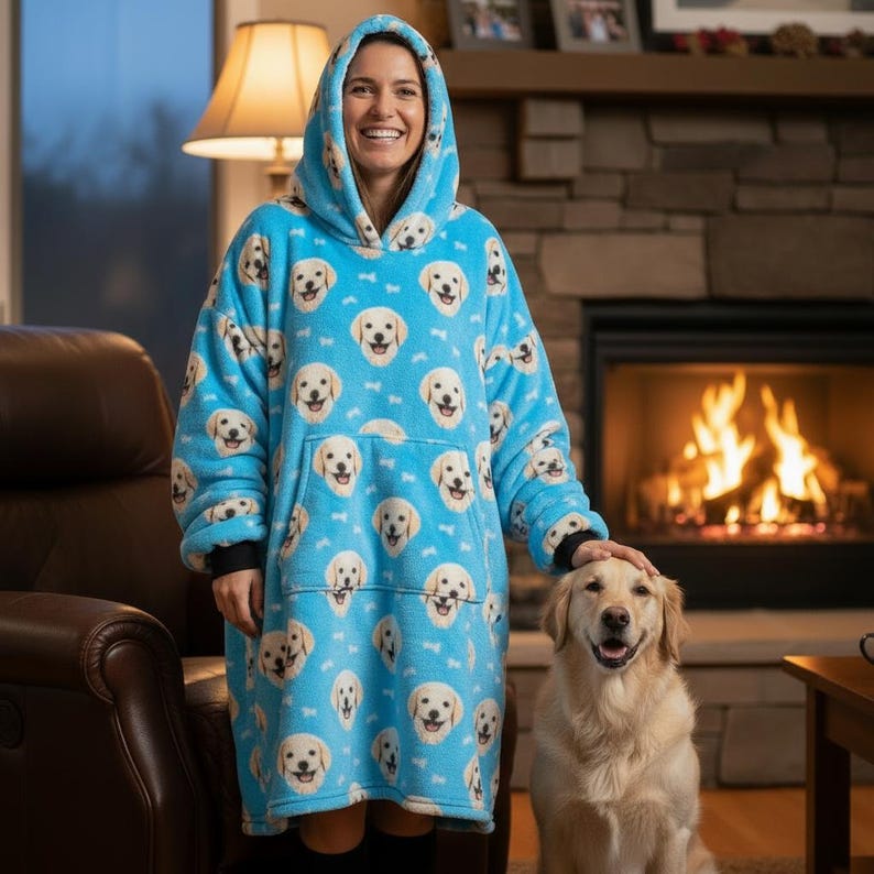 Puede incluir: Una manta con capucha azul claro con un patr&oacute;n repetido de caras de golden retriever y huesos blancos. La manta de gran tama&ntilde;o tiene un gran bolsillo delantero y una capucha. La persona de la imagen lleva la manta.