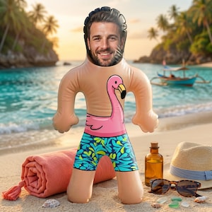 May include: An inflatable doll with a cutout for a face, featuring a printed pink flamingo on the chest and blue and green patterned swim trunks. The doll has a light skin tone and is designed to resemble a person standing on a sandy beach.