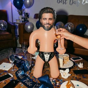 Könnte beinhalten: Aufblasbare Puppe mit männlichem Gesicht und Badeanzug im Smoking-Stil. Die Puppe steht auf einem Tisch mit Sektgläsern, Kuchen und Luftballons. Eine Hand hält ein Sektglas.