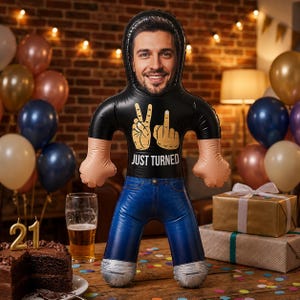 May include: An inflatable balloon shaped like a person with a printed face, wearing a black t-shirt with the text JUST TURNED and hand gestures, and blue jeans. The balloon has tan arms and silver cuffs at the bottom of the pant legs.