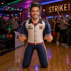 May include: An inflatable figure featuring a printed image of a smiling man's face at the top. The figure wears a cream-colored bowling shirt with a red collar, a crown icon, the text KINGPIN, and a red bowling pin graphic, paired with dark blue pants.