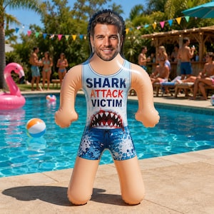 May include: An inflatable pool float shaped like a person with a custom photo face. The float features a light blue tank top with the text SHARK ATTACK VICTIM in blue and red, a graphic of a shark mouth with teeth, and blue patterned swim trunks.