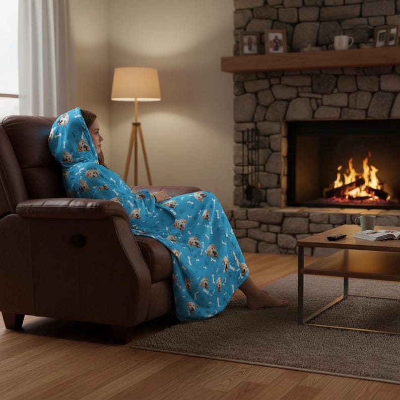 Puede incluir: Una persona est&aacute; sentada en un sill&oacute;n reclinable marr&oacute;n, envuelta en una manta con capucha azul claro. La manta presenta un patr&oacute;n de caras de golden retriever y huesos blancos. Una chimenea con fuego ardiendo est&aacute; en el fondo.