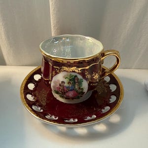 May include: A porcelain teacup and saucer set featuring a deep burgundy glaze with gold trim and accents. The cup and saucer display a central oval medallion depicting two figures in a garden setting, surrounded by intricate gold floral patterns.
