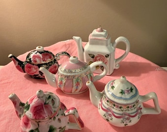 May include: A collection of miniature porcelain teapots in various designs. The teapots feature floral patterns in pink, white, and black, with gold accents. Some have rose motifs, while others have decorative ribbons and trim.