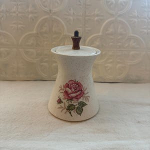 May include: A vintage ceramic sugar bowl with a lid. The off-white bowl has a speckled finish and a floral rose design in pink and green. The lid has a wooden handle. The sugar bowl is a decorative kitchen item.