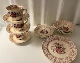 May include: A collection of cream-colored ceramic dinnerware featuring a repeating reddish-brown dotted border pattern and a central floral motif with pink, purple, and green blossoms. The set includes stacked teacups, saucers, and plates.
