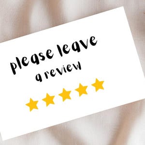 May include: A white card with the text "please leave a review" in black script, and five yellow stars. The card is on a beige fabric surface, with a dried plant stem to the right.