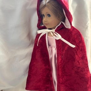 Könnte beinhalten: Eine Puppe mit einem roten Samt-Kapuzenmantel mit weißem Futter und einer weißen Schleife. Die Puppe hat blaue Augen, Sommersprossen und hellbraunes Haar. Die Puppe trägt ein rosa Kleid mit Spitzenbesatz.
