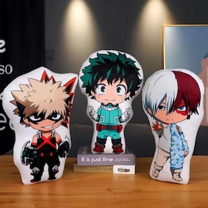 May include: Three character-shaped pillows. One has spiky blonde hair and a black outfit with red accents. The second has green hair and a teal and red outfit. The third has white and red hair and a light blue outfit. The pillows are on a wooden surface.