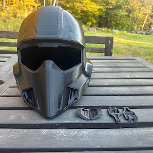 May include: A matte black full-face helmet featuring a prominent angular visor and a pointed chin guard with integrated ventilation ports. Beside the helmet are four separate black plastic components, including two small vents and two frame pieces.