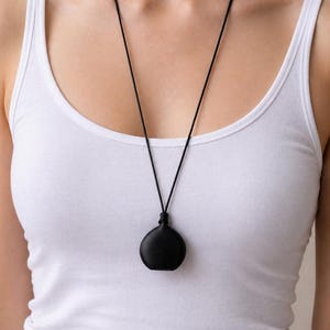 Peut inclure: Un collier avec un pendentif noir en forme de goutte d'eau. Le pendentif est suspendu à un cordon noir fin. La personne porte un débardeur blanc.