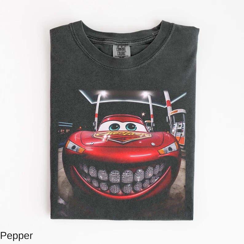 Puede incluir: Camiseta gris oscuro con un gr&aacute;fico de Rayo McQueen con una parrilla de diamantes. La imagen muestra el coche en una gasolinera por la noche. La camiseta est&aacute; doblada y tiene el logo de Comfort Colors.