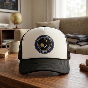 Puede incluir: Gorra de camionero en blanco y negro con un emblema circular. El emblema presenta las palabras "HAIL MARY" y "EARTH" alrededor de un diseño central. La gorra tiene un panel frontal blanco y una parte trasera de malla negra.