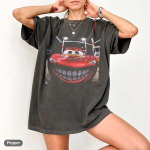 Puede incluir: Camiseta gris oscuro con un gran gr&aacute;fico de un coche de dibujos animados rojo con una amplia sonrisa. El coche est&aacute; ambientado en una escena nocturna con gasolineras. La camiseta tiene mangas cortas y un corte holgado.