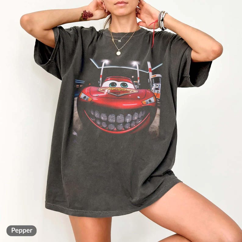 Puede incluir: Camiseta gris oscuro con un gran gr&aacute;fico de un coche de dibujos animados rojo con una amplia sonrisa. El coche est&aacute; ambientado en una escena nocturna con gasolineras. La camiseta tiene mangas cortas y un corte holgado.
