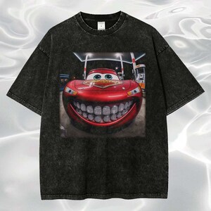 Puede incluir: Camiseta negra con un gr&aacute;fico de un coche de carreras rojo sonriente con una gran parrilla. El coche tiene ojos verdes y est&aacute; ambientado en una gasolinera. La camiseta tiene un aspecto vintage y desgastado.