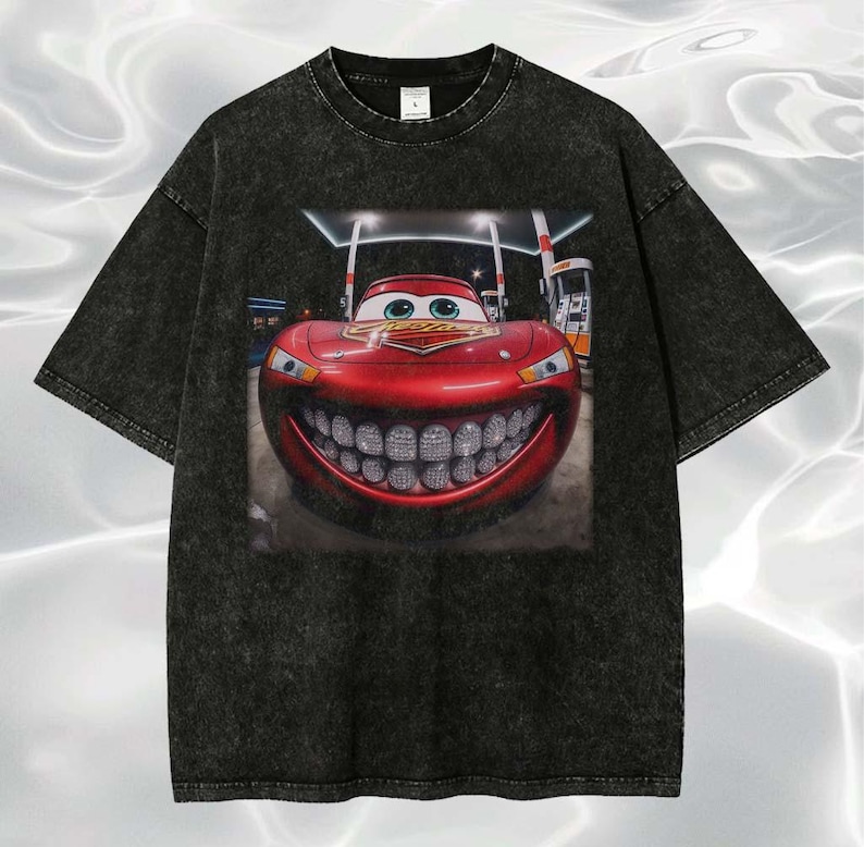 Puede incluir: Camiseta negra con un gr&aacute;fico de un coche de carreras rojo sonriente con una gran parrilla. El coche tiene ojos verdes y est&aacute; ambientado en una gasolinera. La camiseta tiene un aspecto vintage y desgastado.