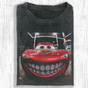 Puede incluir: Camiseta gris oscuro con un gr&aacute;fico de un coche de carreras de dibujos animados rojo con una sonrisa adornada con diamantes. El coche est&aacute; en una gasolinera por la noche. La camiseta tiene cuello redondo y est&aacute; doblada.