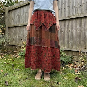 May include: A long, patchwork skirt in shades of red, brown, and burgundy. The skirt features a floral pattern and a tiered design. The model is wearing the skirt with a light blue t-shirt and brown shoes, standing in a grassy area.