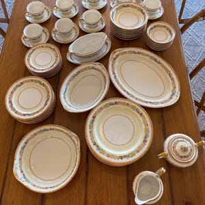 May include: A complete vintage tea set with floral and gold trim. The set includes teacups and saucers, serving dishes, and a teapot and creamer. The dishes are arranged on a wooden table.