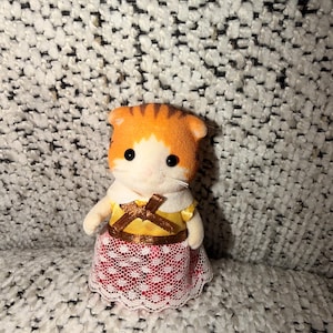 Puede incluir: Una peque&ntilde;a figura de gato naranja y blanco, vestida con un vestido amarillo con un lazo marr&oacute;n y una falda de encaje rojo y blanco. El gato tiene ojos negros y un cuello blanco.
