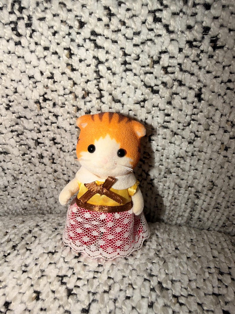 Puede incluir: Una peque&ntilde;a figura de gato naranja y blanco, vestida con un vestido amarillo con un lazo marr&oacute;n y una falda de encaje rojo y blanco. El gato tiene ojos negros y un cuello blanco.