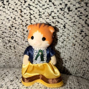 Puede incluir: Una figura en miniatura de un gato naranja y blanco que lleva una chaqueta azul marino con ribete de encaje blanco y una falda amarilla con detalles de cinta marr&oacute;n. El gato tiene ojos negros y hocico blanco.