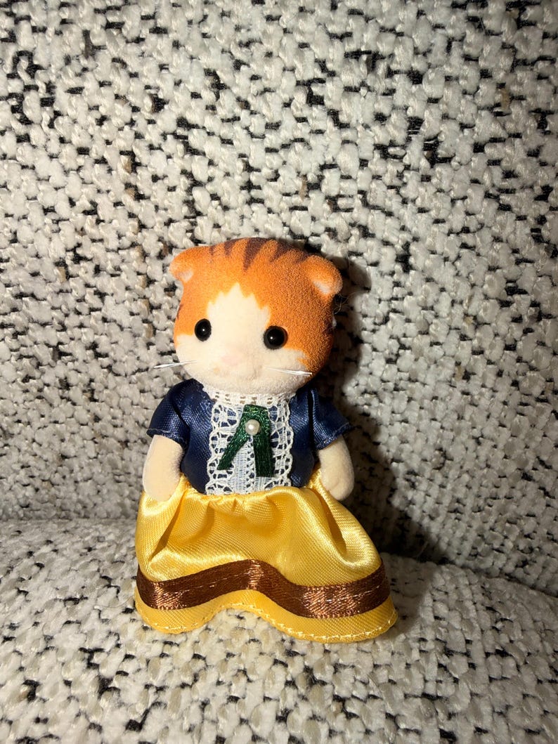 Puede incluir: Una figura en miniatura de un gato naranja y blanco que lleva una chaqueta azul marino con ribete de encaje blanco y una falda amarilla con detalles de cinta marr&oacute;n. El gato tiene ojos negros y hocico blanco.