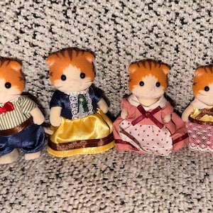 Puede incluir: Cuatro figuras de gatos en miniatura con pelaje naranja y blanco, cada una vestida con diferentes atuendos. Uno lleva una camisa a rayas y pantalones azules, otro un vestido azul con una falda amarilla, y los otros visten vestidos rosas.