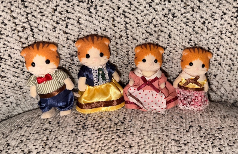 Puede incluir: Cuatro figuras de gatos en miniatura con pelaje naranja y blanco, cada una vestida con diferentes atuendos. Uno lleva una camisa a rayas y pantalones azules, otro un vestido azul con una falda amarilla, y los otros visten vestidos rosas.