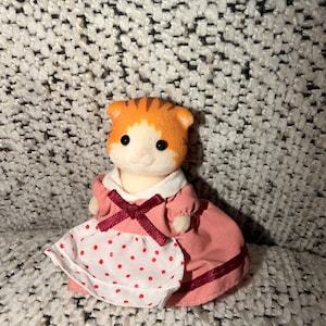 Puede incluir: Una peque&ntilde;a figura de gato naranja y blanco con un vestido rosa con un delantal blanco con lunares rojos. El vestido tiene un cuello blanco y un lazo burdeos. El gato tiene ojos negros y una cabeza rayada.
