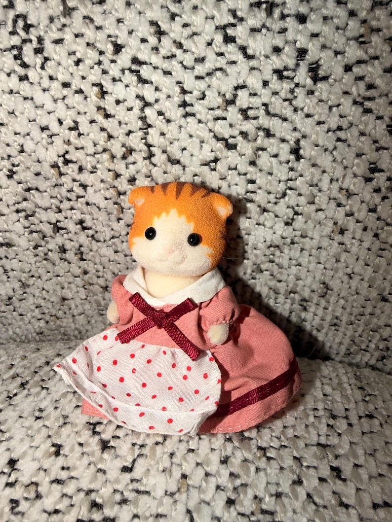 Puede incluir: Una peque&ntilde;a figura de gato naranja y blanco con un vestido rosa con un delantal blanco con lunares rojos. El vestido tiene un cuello blanco y un lazo burdeos. El gato tiene ojos negros y una cabeza rayada.